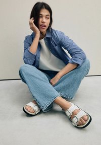 Sandali bianchi a due cinturini con fibbie, indossati con jeans di jeans a gamba larga di un azzurro chiaro e una camicia a righe blu sopra una t-shirt bianca.