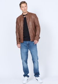 Braune Lederjacke mit abgesteppten Schultern, Reißverschluss vorne, kombiniert mit einem schwarzen T-Shirt und hellblauen Jeans. Weiße Sneakers runden das Outfit ab.