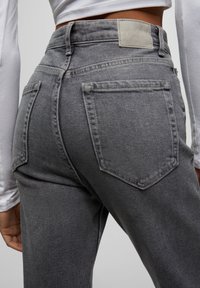 Le jean en denim gris présente une taille haute, un design à cinq poches et un tissu texturé. L'arrière comprend une étiquette en cuir avec le logo de la marque.