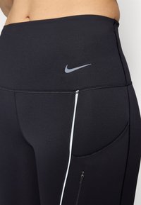 Crne sportske tajice s glatkom tkaninom, visokim strukom, sivoj Nike oznaci i bočnim džepom sa zatvaračem i bijelim detaljem.