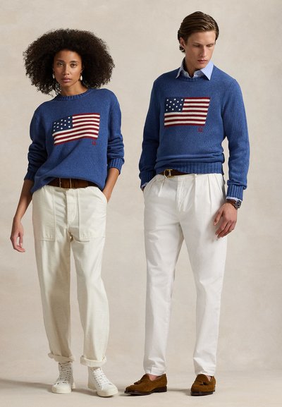 Zwei Models tragen passende blaue Pullover mit amerikanischen Flaggenmotiven, kombiniert mit weißen Hosen und braunen Gürteln, und stehen vor einem schlichten Hintergrund.