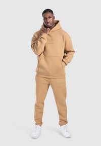Hombre de pie con la mano cerca de la barbilla, vistiendo una sudadera beige y pantalones a juego con zapatillas blancas, frente a un fondo liso.