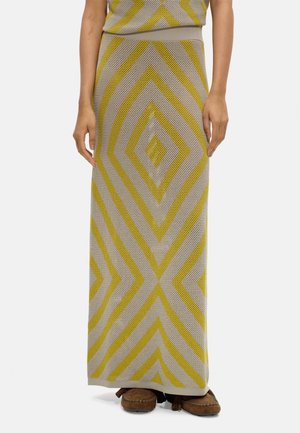 Długa beżowa dzianinowa spódnica z żółtym wzorem diamentu, noszona z brązowymi mokasynami, pokazująca dolną część tułowia i nogi.