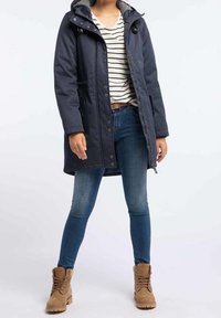 Parka azul marino con capucha, botones frontales y bolsillos laterales, llevada sobre una camisa a rayas y jeans azules. Botas marrones con cordones completan el atuendo.