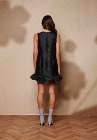 Μαύρο αμάνικο φόρεμα με βολάν στο τελείωμα, με υφή σχεδίου. Η μοντέλο το συνδύασε με ανοιχτό γκρι καλτσάκια και τακούνια πάνω σε ξύλινο πάτωμα.