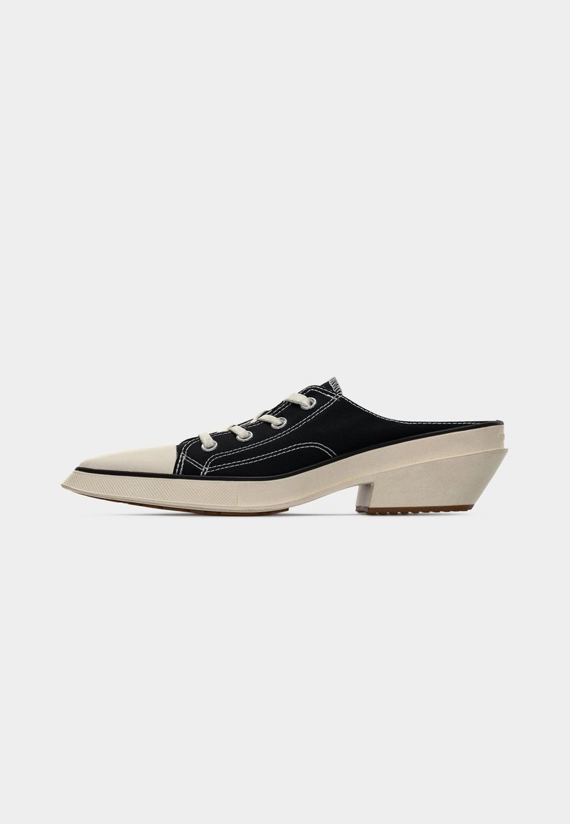 Sneaker mule nera e beige con lacci bianchi, cuciture bianche e tacco a zeppa basso su uno sfondo semplice.