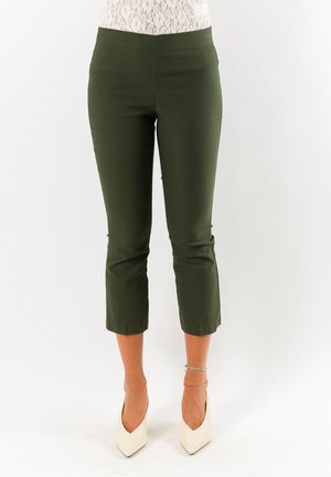 Donna con pantaloni corti verde oliva, tacchi alti bianchi a punta e braccialetti alla caviglia, in piedi su sfondo bianco.