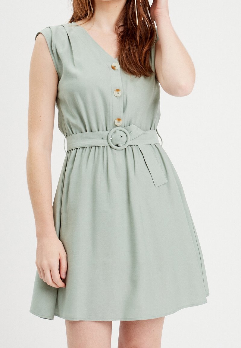 Robe vert clair en tissu doux, avec un col en V, des boutons sur le devant et une ceinture avec une boucle ronde. Style avec des manches courtes et une jupe évasée.