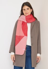 Een roze en rode sjaal met een patroon, gedragen over een lichte trui en een grijze jas, met een zachte textuur en een geometrisch ontwerp in verschillende tinten.