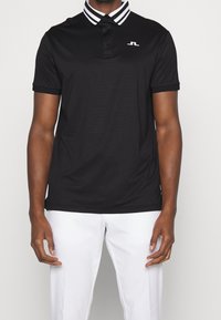 Svart polo t-shirt med vit och svart randig krage, korta ärmar och en liten logotyp på bröstet. Tillverkad av andningsbart material.