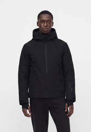 Homme portant une veste noire à capuche avec fermeture éclair et trous pour les pouces dans les manches, debout devant un fond uni gris clair.