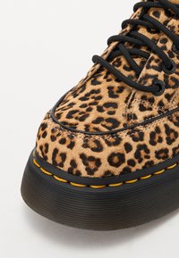 Zapatillas de leopardo con un upper texturizado en marrón claro, cordones negros y una suela de goma negra gruesa con un acento de costura amarilla.