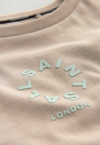Ljus beige bomullströja med rundad hals, prydd med upphöjd mintgrön text som säger "SAINT LAC" och "LONDON" i en cirkulär design.