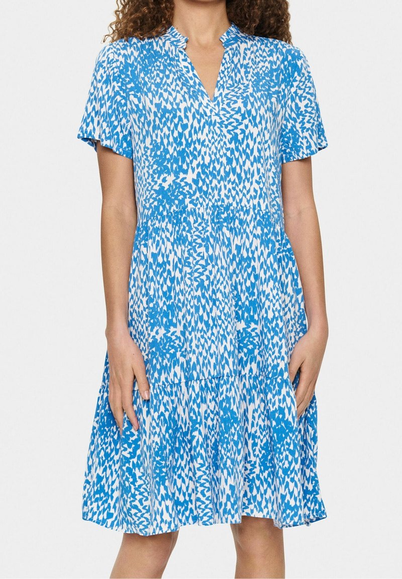 Femme portant une robe courte à manches courtes, longueur genoux, avec un motif floral abstrait bleu et blanc et un ourlet froncé sur fond uni.