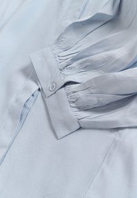 Chemise bleu clair avec un col boutonné, des détails de manches plissées et une texture lisse. Le tissu semble léger et aéré.