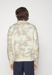 Felpa mimetica beige con scollo rotondo, polsini e orlo a costine. Presenta una texture liscia e una tasca sulla manica sinistra.