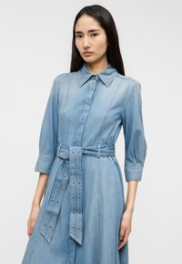 Φόρεμα από ανοιχτό μπλε τζιν με γιακά, μανίκια έως τον αγκώνα και ζώνη στη μέση. Διαθέτει κλείσιμο με κουμπιά και κάθετες ραφές.