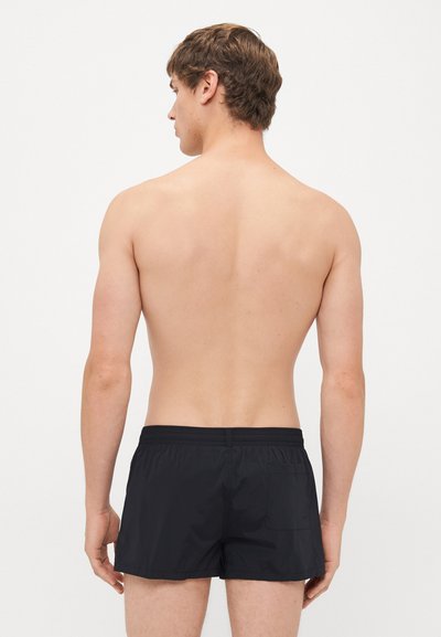 Ung man som vänder ryggen mot kameran, iförd svarta badbyxor med elastisk midja och en liten bakficka, mot en vit bakgrund.