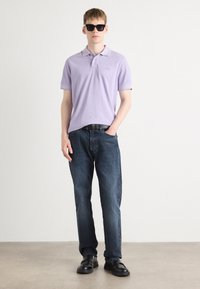 Lilla polo skjorte med korte ærmer, mørkeblå jeans og sorte sko. Modellen bærer svarte solbriller; har en diskret logo detalje på skjorten.