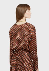 Pitkähihainen bleiseri ruskeasta silkistä, jossa on geometrinen kuvio. Pyöreä kaula-aukko ja rento istuvuus, jossa on kontrastiväreinä beigejä ja mustia muotoja.