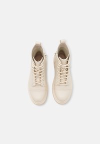 Une paire de baskets montantes beiges avec lacets, fermeture éclair latérale et languettes, présentée de dessus sur un fond blanc uni.
