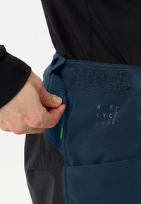 Donkerblauwe stoffen tailleband met klittenbandsluiting; beschikt over een logoafdruk en een groene tag. Handmatig aanpassen van de sluiting is zichtbaar.