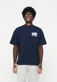 Mørkeblå bomulds t-shirt med korte ærmer og rund hals. Har et logo på venstre bryst. Båret med lysegrå sweatpants.