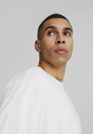 Fehér, oversized póló környakkal, lehulló vállakkal. Finom textúrájú, a nyakon látható "BLESSED" tetoválás nyomtatva.