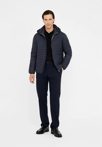 Veste matelassée bleu marine à capuche, texture côtelée, fermeture éclair et poches latérales, associée à un pantalon foncé et des chaussures noires.