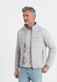 Lys grå pufferjakke med vandret syninger, høj krave og lynlås-lommer, båret over en lys grå sweater og blå jeans.