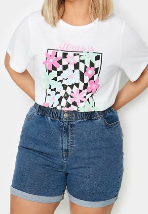 Femme portant un t-shirt blanc avec des fleurs pastel et un imprimé à carreaux noirs, associé à un short en jean bleu à ourlet roulé.