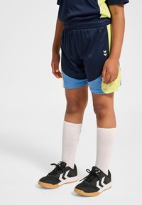 Mörkblå sportshorts med gröna och blå accenter, utrustade med dragsko i midjan. Svarta skor med vita accenter och vita strumpor.