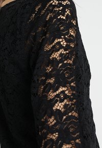 Manica in pizzo floreale nero su pelle chiara, che mostra un dettagliato motivo trasparente e una texture raffinata.