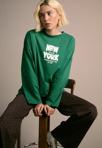 Zelena košulja dugih rukava s bijelim grafičkim natpisom "NEW YORK"; uparen s smešnim hlačama s leopard uzorkom. Model sjedi na drvenoj stolici, ruku preklopljenih.