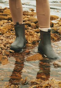 Zielone wodoodporne buty z gładkim wykończeniem, brązowymi akcentami, niską wysokością cholewki i uchwytem do zakupu, stojące w płytkiej wodzie na skalistym terenie.