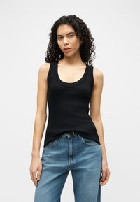 Femeie cu păr creț, purtând un top negru fără mâneci, mulat, băgat în blugi albaștri cu talie înaltă, stând în picioare pe un fundal simplu, deschis la culoare.