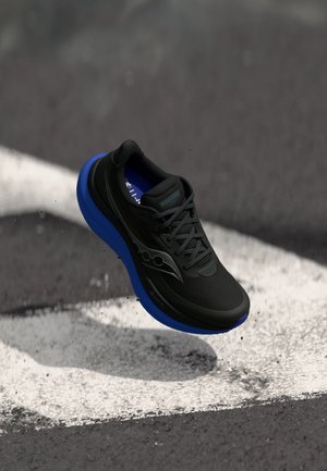 Chaussure de course noire et bleue avec des lacets, en l'air au-dessus d'un asfalt avec des lignes blanches peintes, petits débris éparpillés autour.