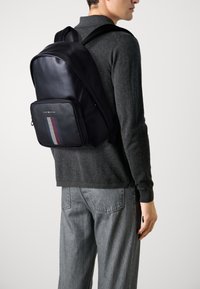 Zaino in pelle nera con texture liscia, design snodato, tasca frontale con zip caratterizzata da un motivo a strisce rosso, bianco e blu.