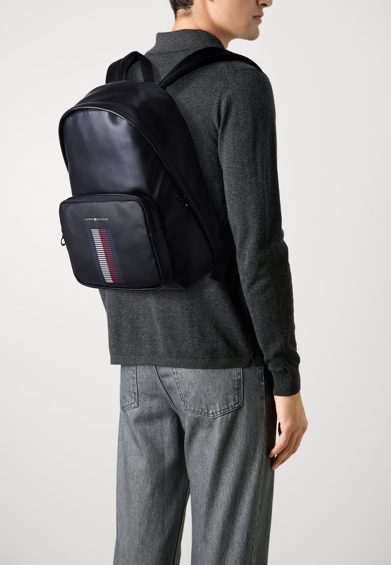 Zaino in pelle nera con texture liscia, design snodato, tasca frontale con zip caratterizzata da un motivo a strisce rosso, bianco e blu.