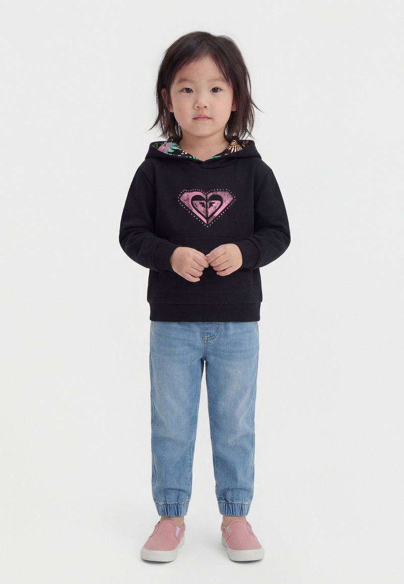 Νεαρό παιδί όρθιο, φοράει μαύρο φούτερ με κουκούλα και ροζ λογότυπο καρδιάς, ανοιχτόχρωμα μπλε τζιν και ροζ αθλητικά χωρίς κορδόνια σε λευκό φόντο.