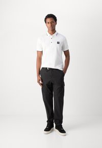 Maglietta polo bianca con bordi neri ed emblema, abbinata a pantaloni cargo neri e scarpe da ginnastica nere con suola chiara. Uomo in piedi, mani nelle tasche.