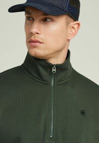 Chaqueta de cremallera verde oscura con cuello alto, tejido texturizado y pequeño logo negro. Se lleva con una gorra de mezclilla oscura que tiene la parte trasera de malla.