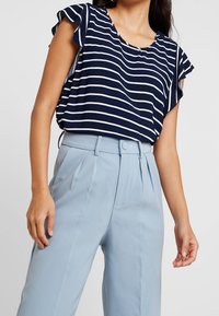 Top a righe navy e bianche con maniche a volant, abbinato a pantaloni a vita alta azzurri chiari con pieghe e chiusura con bottone.
