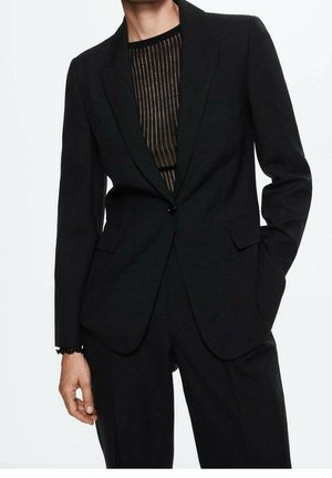 Personne portant un blazer noir sur mesure et un pantalon noir assorti, par-dessus un haut noir transparent à rayures verticales, une main dans la poche.