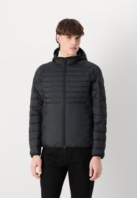 Svart puffjacka med dragkedja fram, huva och ribbade muddars. Har horisontellt kviltad design och en patch-ficka på vänster arm.