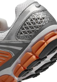 Men's athletic shoe featuring a mesh upper in white with silver accents, a patterned gray heel, and an orange cushioning sole.