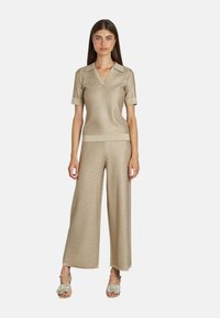 Femme portant un haut coll rond en maille texturée or à manches courtes et un pantalon large assorti, avec des talons ouverts embellis, se tenant droite.
