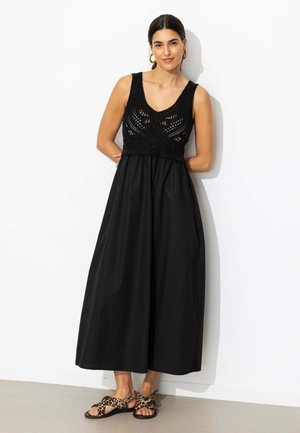 Femme portant une robe noire sans manches en crochet et des sandales à imprimé léopard, debout contre un mur blanc les mains dans le dos.