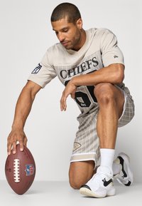 Μπεζ φανέλα NFL Chiefs με διχτυωτές λεπτομέρειες, γκρι σορτς με κάθετες ρίγες και λευκά sneakers με μαύρες λεπτομέρειες. Κρατώντας μια μπάλα του φούτμπολ.