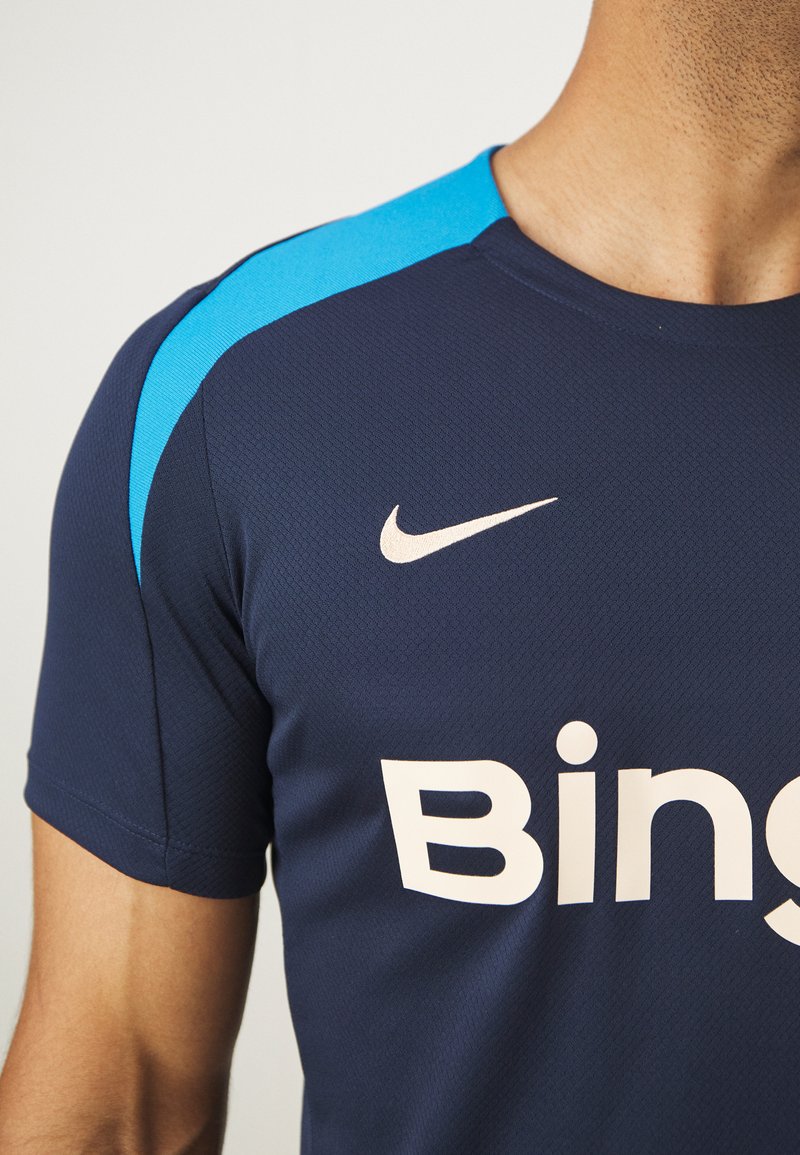 Granatowa koszulka Nike z jasnoniebieskimi akcentami na ramionach. Posiada duże białe logo "Bing" oraz mały biały swoosh. Teksturowany materiał.